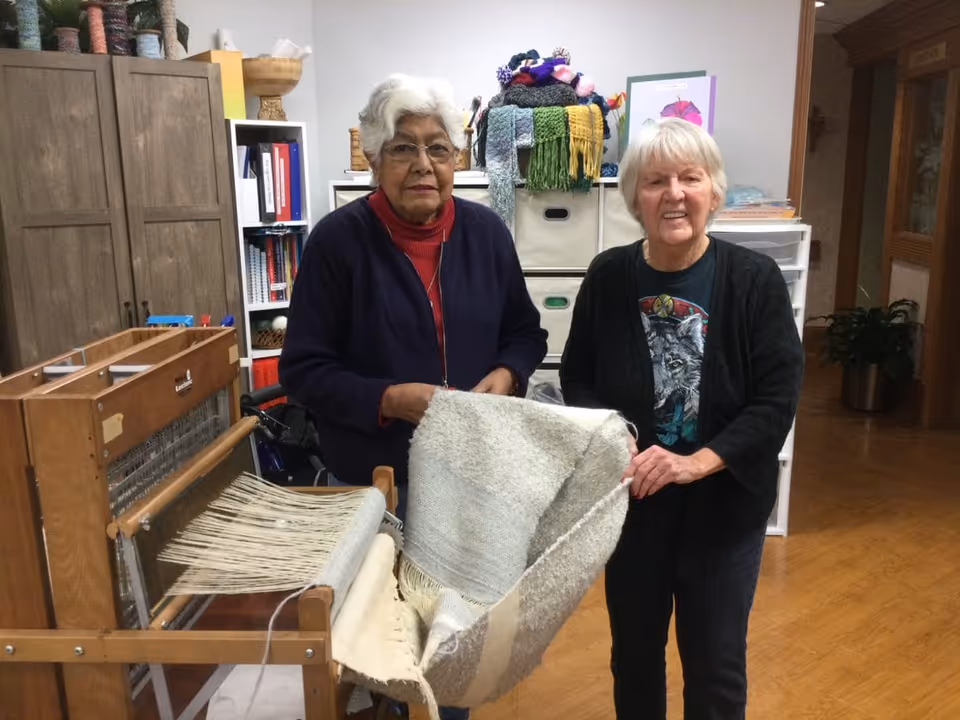 Two elderly women standing indoors next to a wooden weaving loom, holding a partially woven textile. Behind them are shelves with colorful yarns, books, and storage bins. The room has wooden flooring and a cozy, organized atmosphere.