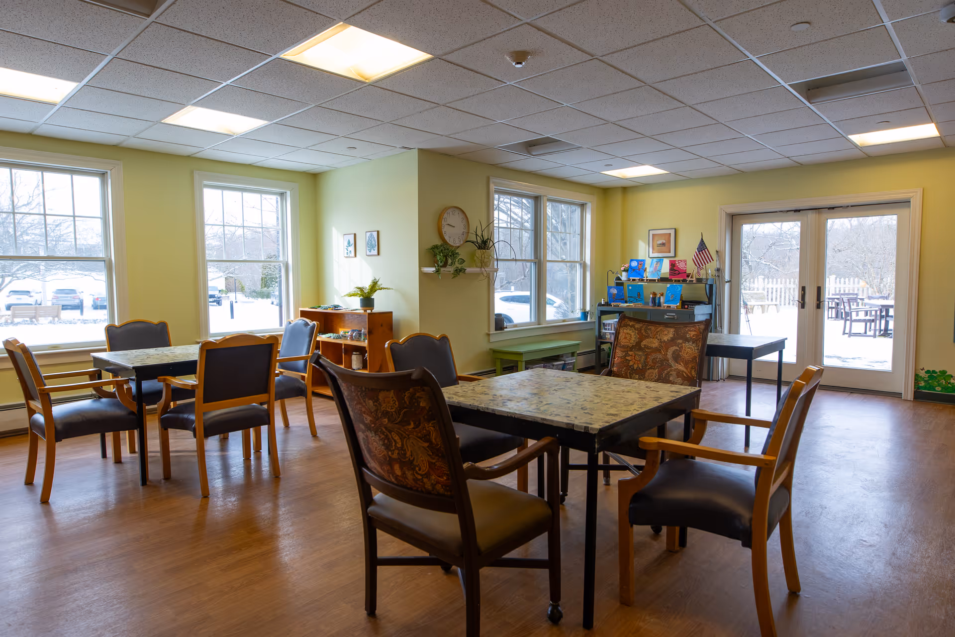 A bright and spacious dining area with several tables and chairs arranged for seating. The room has large windows letting in natural light, light yellow walls, and a wooden floor. There are some plants, a clock on the wall, and a door leading to an outdoor patio area with tables and chairs visible through the glass.
