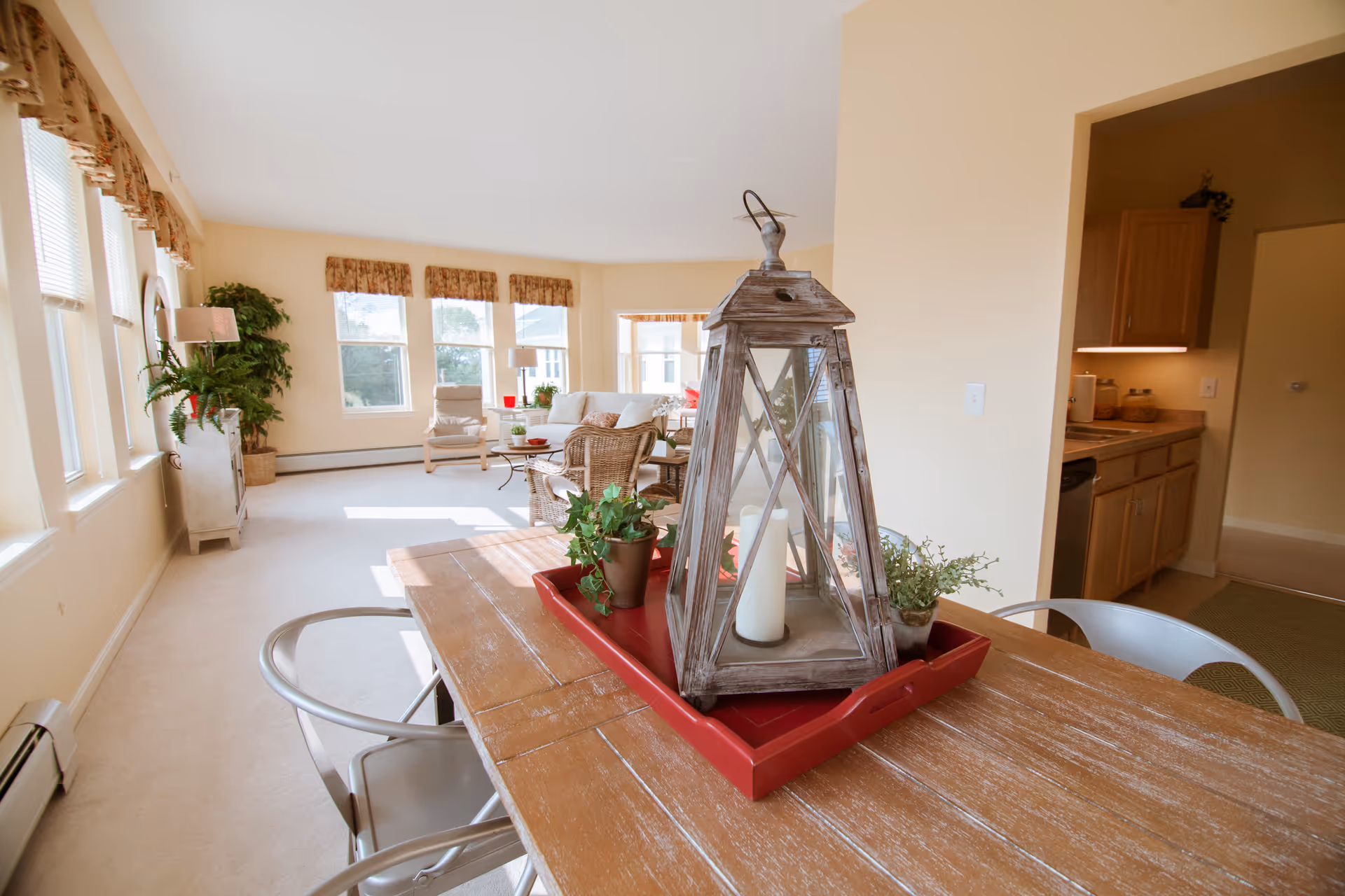 Bright and spacious living area with large windows letting in natural light. In the foreground, a wooden table with a red tray holding a decorative lantern and small potted plants. The background shows a cozy seating area with armchairs, a sofa, and side tables with lamps and plants. To the right, a partial view of a kitchen with wooden cabinets and countertop appliances.