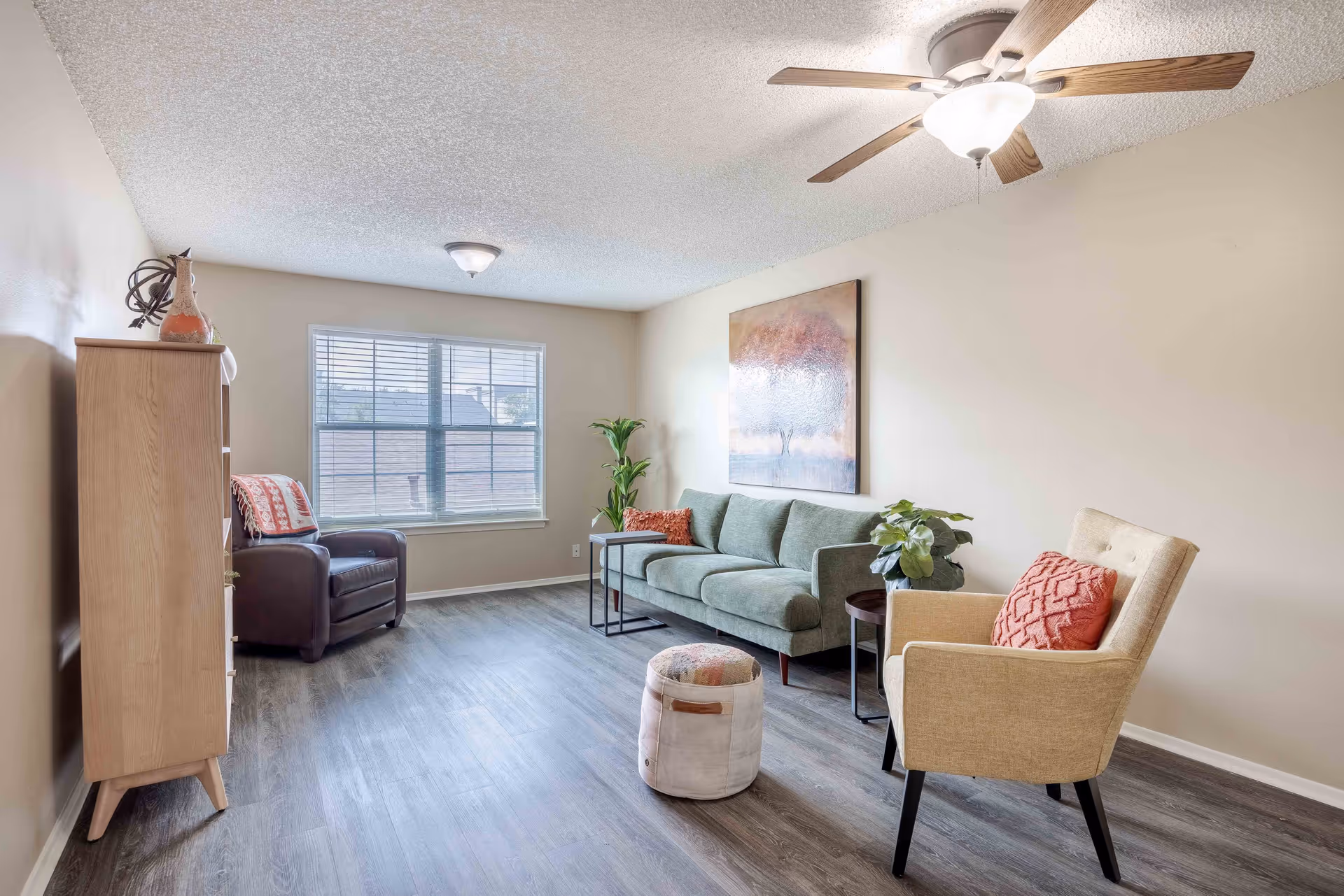 A bright living room with a large window, a green sofa with an orange pillow, a beige armchair with an orange pillow, a small round ottoman, a wooden cabinet, a brown leather armchair with a blanket, two side tables with plants, a ceiling fan with light, and a painting of a tree on the wall.