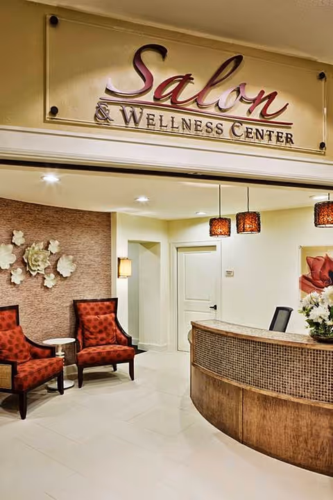 Reception area of a Salon & Wellness Center with a curved wooden reception desk, three hanging pendant lights, two red patterned armchairs, a small round side table, and decorative wall flowers.