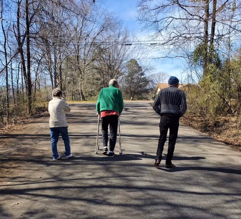 Three elderly individuals walking on a paved road surrounded by leafless trees and some greenery under a clear blue sky. One person is using a walker, while the other two walk alongside.