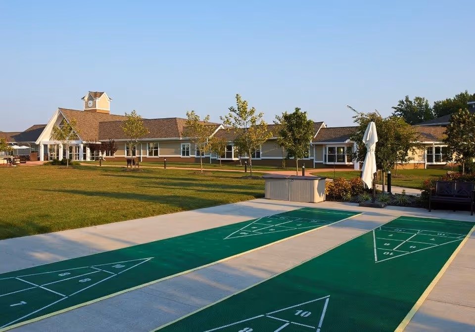 Outdoor area of a senior living facility with two shuffleboard courts in the foreground, a well-maintained lawn, young trees, and a single-story building with a clock tower in the background under a clear blue sky.
