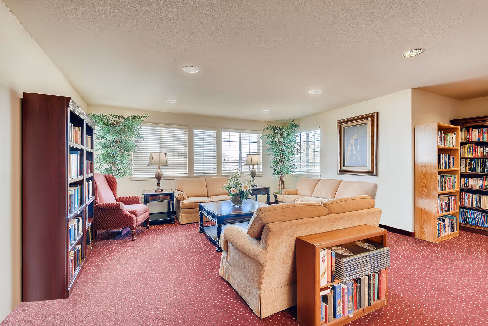 A cozy living room area with beige sofas arranged around a dark wooden coffee table with a flower arrangement. The room has red carpet flooring, large windows with blinds, two table lamps, two tall green plants, a red armchair, and multiple bookshelves filled with books. A framed picture hangs on the wall.