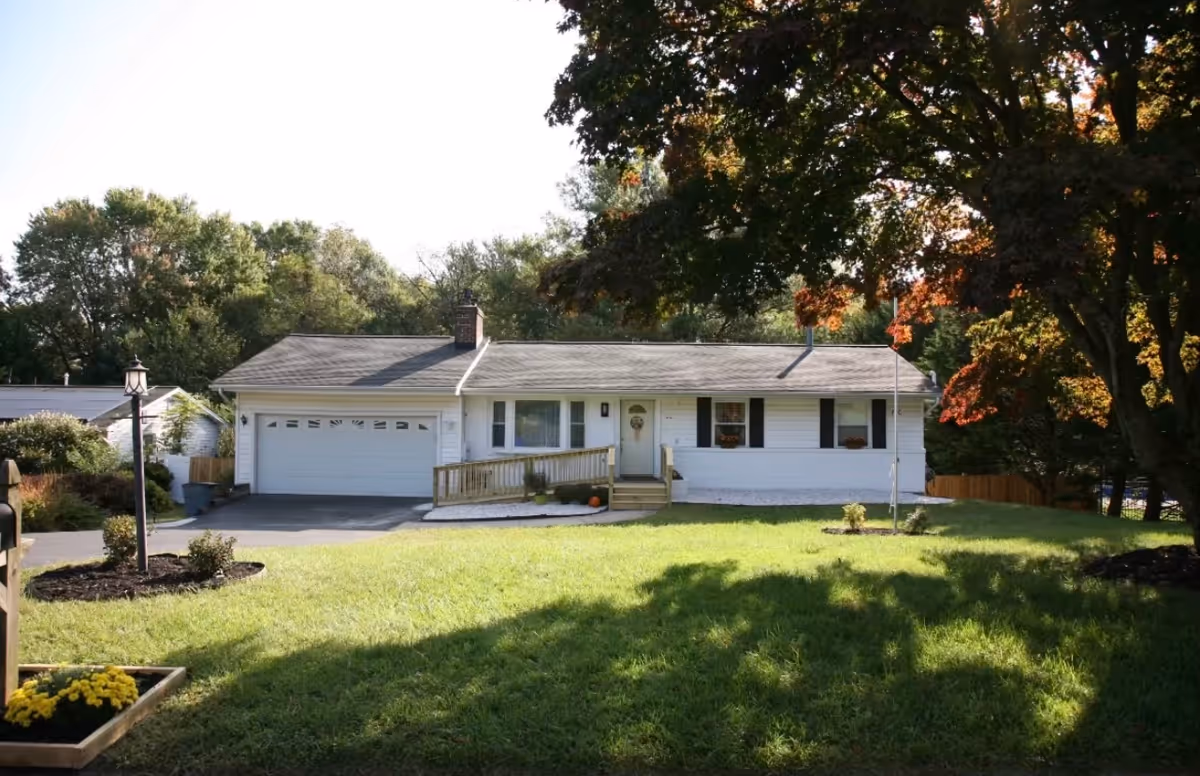 Front exterior of a single-story white ranch-style house with an attached garage, ramped entrance, and a grassy front yard shaded by trees.