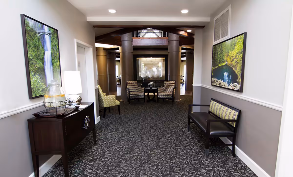 A hallway in a senior living facility with patterned carpet, two framed nature photographs on the walls, a dark wooden console table with a lamp and a glass beverage dispenser, and seating including a bench with a cushion and several armchairs near large columns and a glass partition.