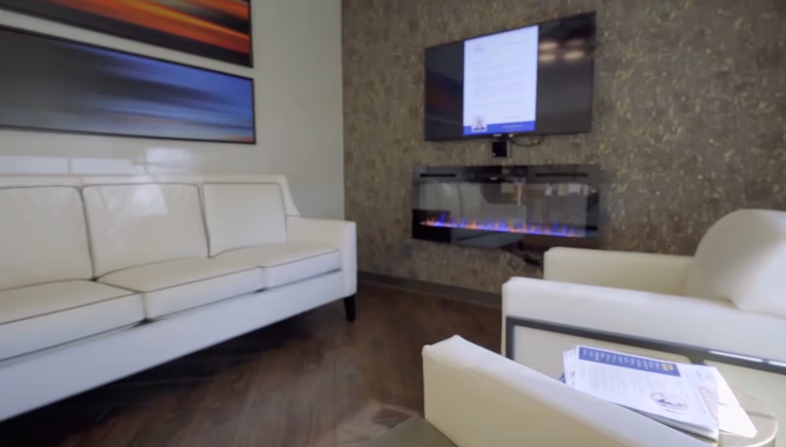 A modern living room with white leather sofas, a wall-mounted flat screen TV above a sleek electric fireplace, and abstract artwork on the wall.