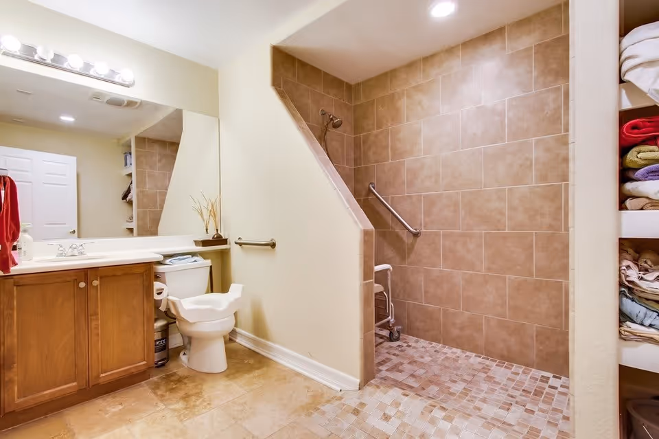 A bathroom with a wooden vanity and sink on the left, a toilet with a raised seat in the center, and a walk-in tiled shower with a grab bar and a shower chair on the right. There are shelves with folded towels visible on the far right side.
