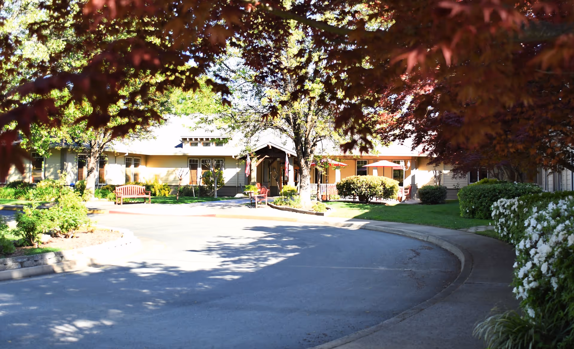View of the front exterior of Oakdale Heights Assisted Living facility with a curved driveway, benches, trees with red and green leaves, bushes, and a building entrance in the background.