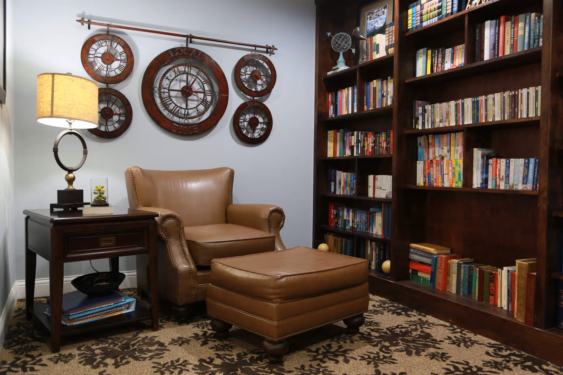 Cozy reading nook with a leather armchair and ottoman beside a side table lamp and a large bookshelf filled with books.