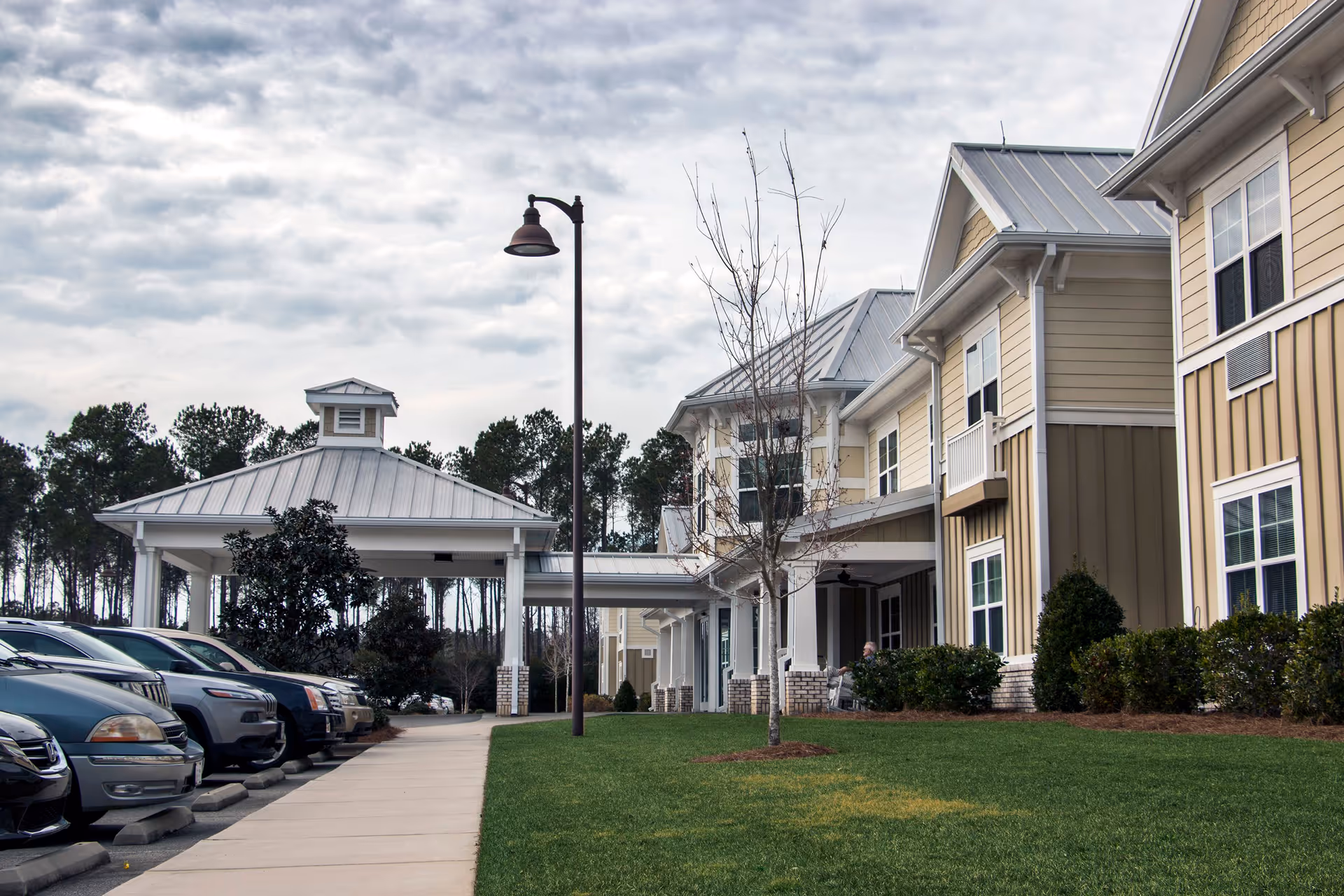 Exterior view of Arbor Landing at Ocean Isle showing a covered entrance with a metal roof, a sidewalk leading to the entrance, parked cars on the left, and a grassy area with a small tree on the right. The building has beige and brown siding with white trim and multiple windows.