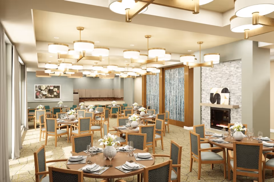 A spacious dining room with multiple round tables set with plates, napkins, glasses, and floral centerpieces. The room features modern light fixtures hanging from the ceiling, large windows with curtains on one side, and a decorative fireplace with abstract art above it on the other side. The walls are painted in soft neutral tones with wood accents and a patterned carpet covers the floor.