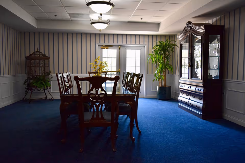 A dining room with a dark wooden dining table and six matching chairs on a blue carpet. The room has striped wallpaper, a glass-fronted wooden cabinet on the right, a large potted plant near the back door, and a decorative birdcage with a plant on the left. Two ceiling lights illuminate the space.