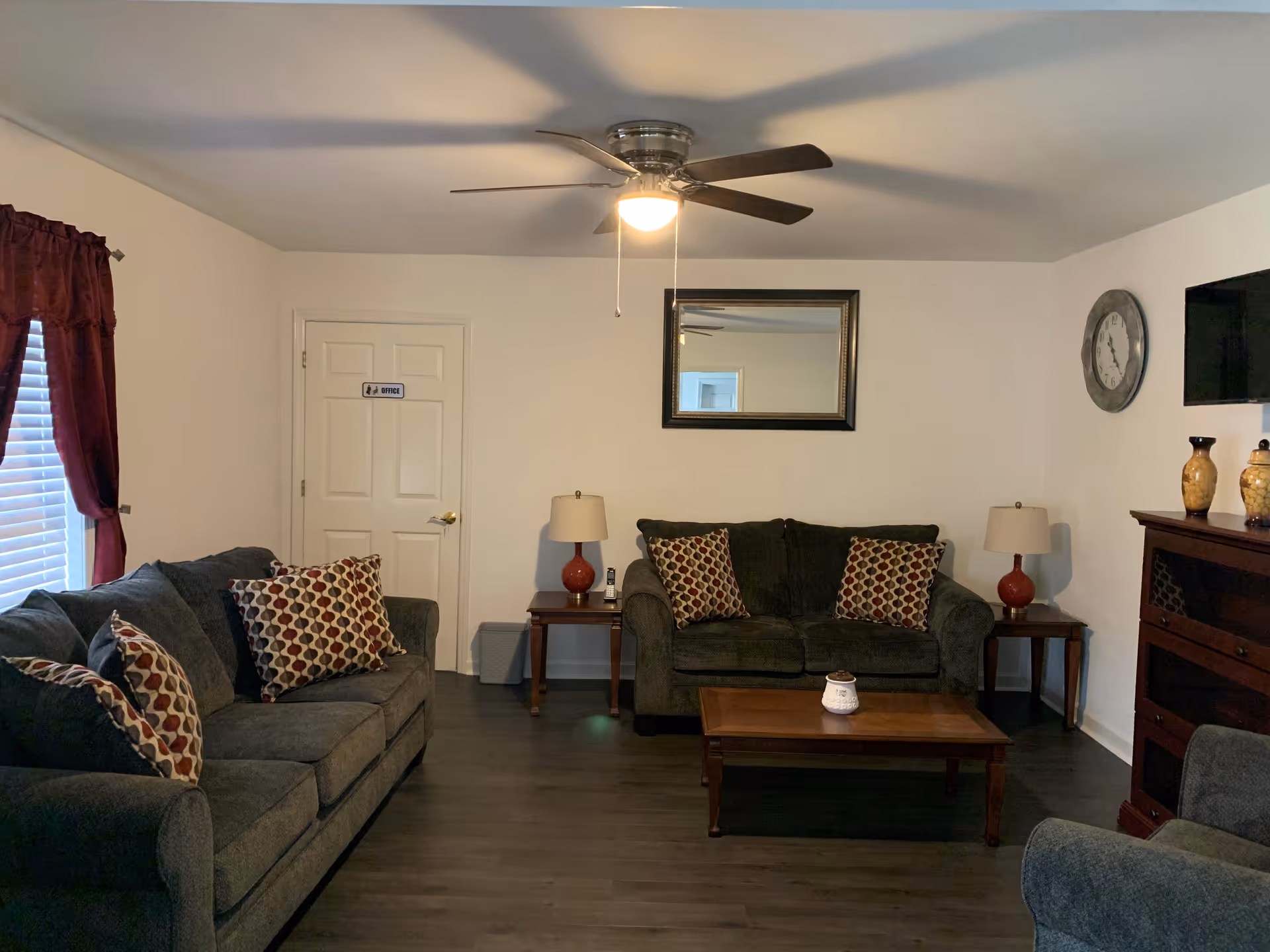 A cozy living room with two dark gray sofas adorned with patterned pillows, a wooden coffee table in the center, two side tables with matching lamps, a wall-mounted TV, a large mirror, a wall clock, and a ceiling fan with a light. There is a door labeled 'Office' and a window with red curtains.