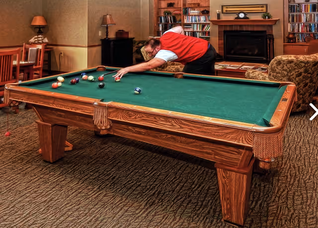 A man in a red vest and white shirt is playing pool on a green felt pool table in a cozy room with carpeted floor. The room has a fireplace with a clock on the mantle, bookshelves filled with books, a patterned sofa, and a wooden dining table with chairs. Two table lamps provide warm lighting.