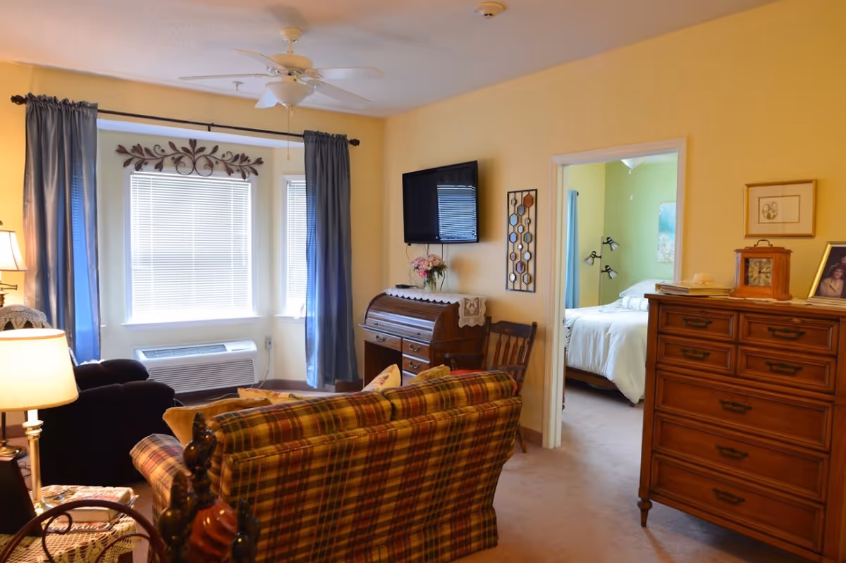 Cozy living room with a plaid sofa, armchair, wall-mounted TV, and an open doorway leading to a bedroom.