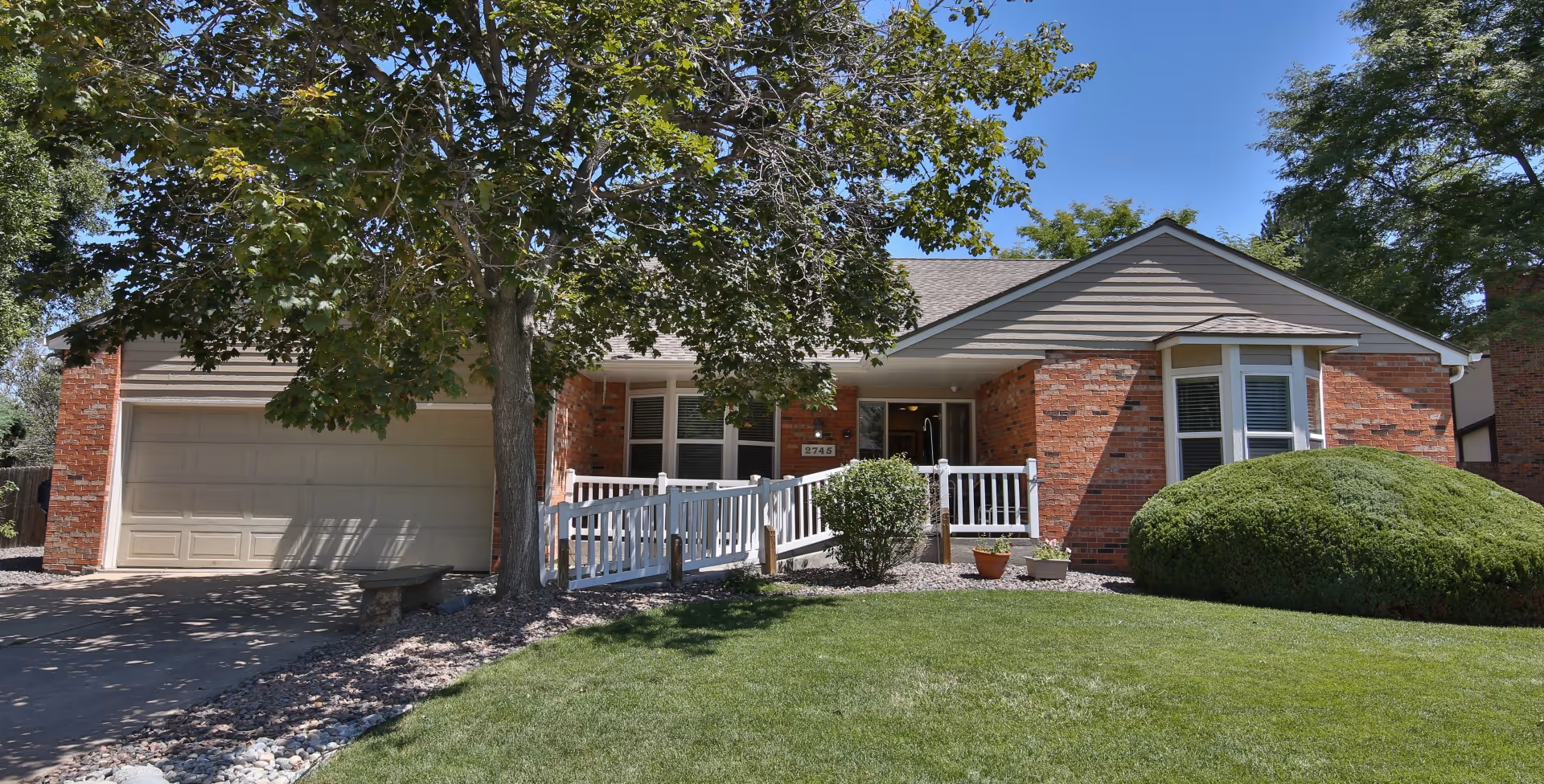 Front exterior of a single-story brick building with a garage, white porch ramp, trees, and a manicured lawn.