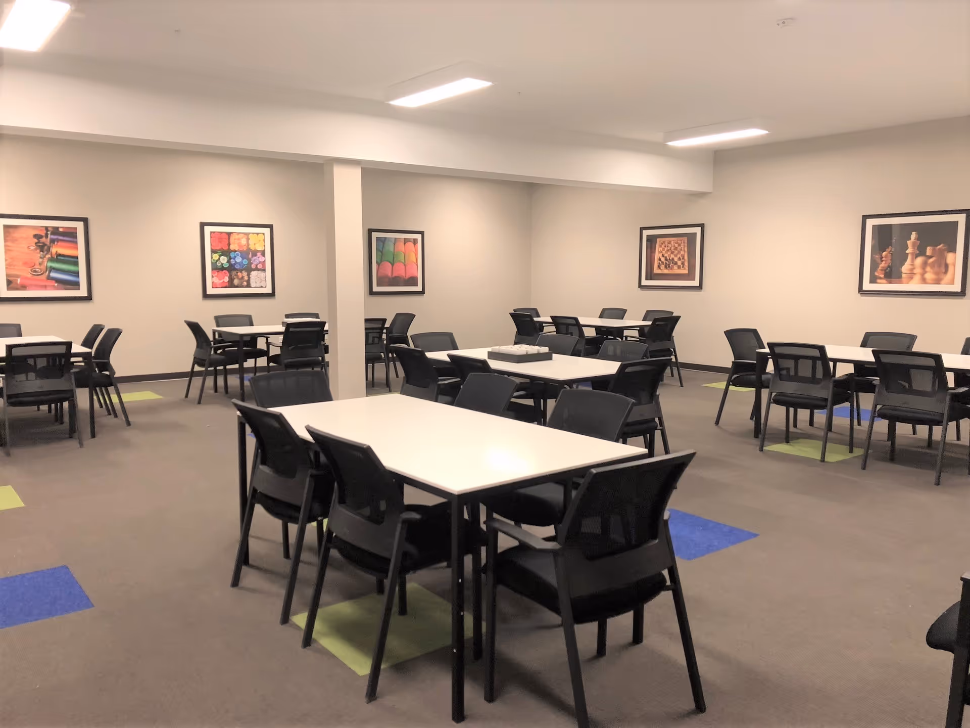 A well-lit room with multiple white tables and black chairs arranged neatly. The carpet has colorful square patterns in blue and green. The walls are decorated with framed pictures featuring chess pieces and colorful objects.
