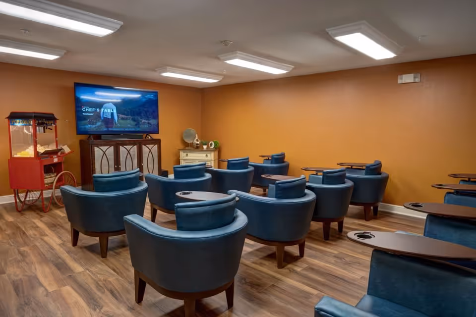A small theater room with multiple blue armchairs arranged in rows facing a large flat-screen TV mounted on an orange wall. There is a red popcorn machine on the left side of the room and a small cabinet with decorative items beneath the TV. The floor is wood and the ceiling has bright fluorescent lights.