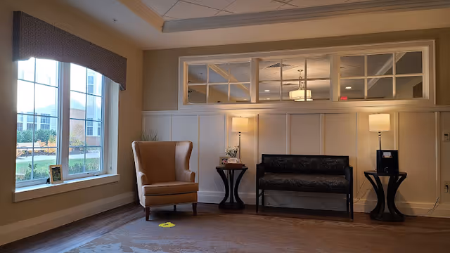 A cozy seating area with an armchair, bench, side tables and lamps beside a large window in a nursing facility common room.