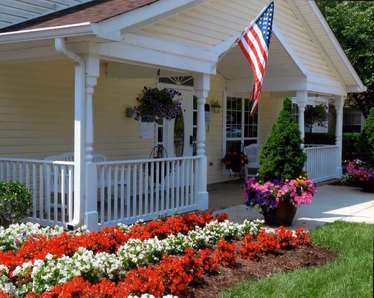 Front porch of a building with white railings and columns, an American flag hanging from the roof, hanging flower baskets, a potted plant with colorful flowers, and a well-maintained flower bed with red and white flowers in front.