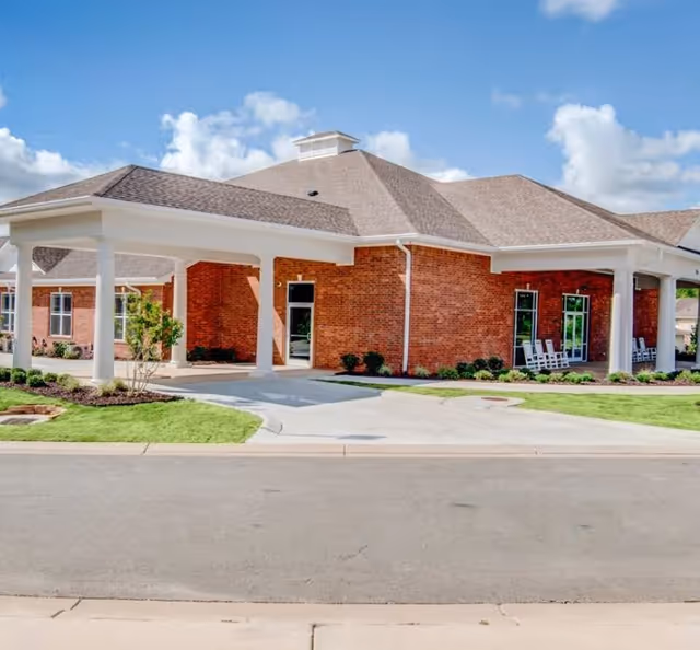 Exterior view of a single-story brick building with a covered entrance supported by white columns, surrounded by neatly maintained grass and small shrubs under a partly cloudy blue sky.