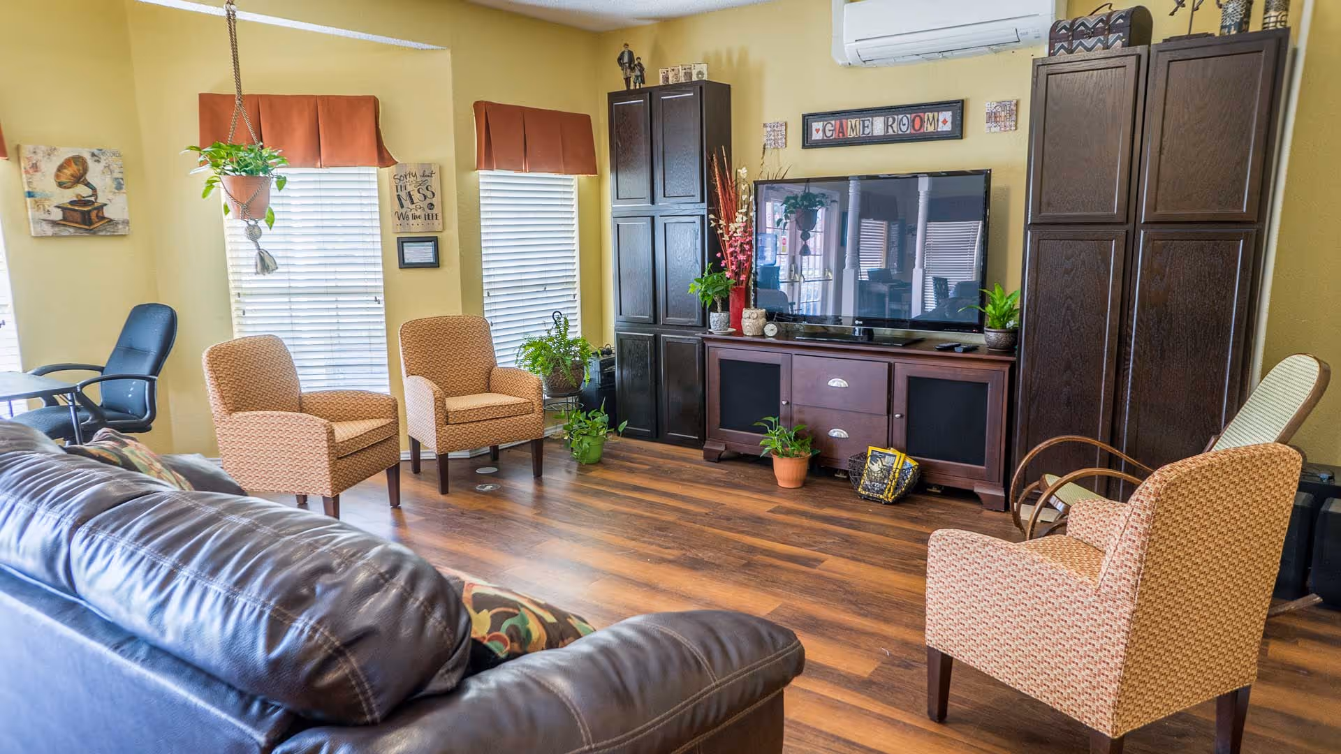 A cozy living room with a dark leather couch, three patterned armchairs, a large flat-screen TV on a wooden cabinet, and several potted plants. The walls are painted yellow and decorated with framed art and a 'Game Room' sign above the TV. There are two windows with blinds and red valances, and a hanging plant near the windows.