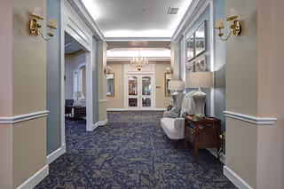 A well-lit hallway in a senior living facility with patterned blue carpet, beige walls, and white trim. The hallway features wall sconces with lampshades, a small upholstered bench with decorative pillows, a wooden side table with a lamp, and framed artwork on the walls. At the end of the hallway, there are double glass doors with a chandelier hanging above.