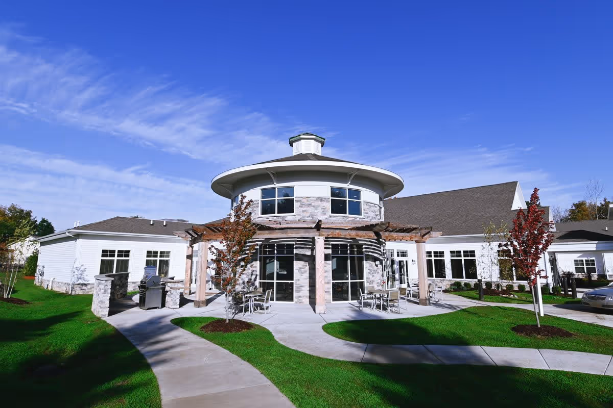 Exterior view of a senior living facility building with a round central section featuring large windows and a wooden pergola. There are outdoor tables and chairs on a concrete patio surrounded by green grass and young trees under a clear blue sky.