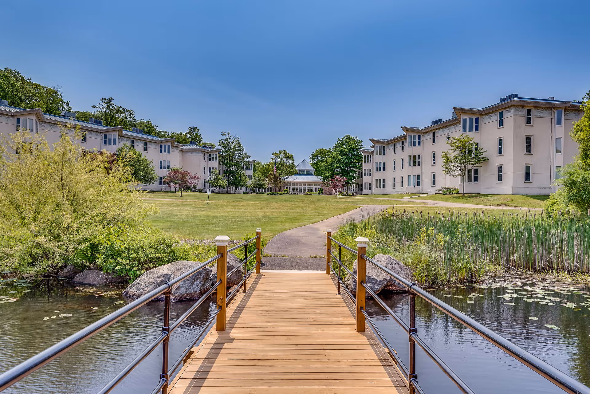 A wooden footbridge over a pond leads to a grassy courtyard framed by multi-story white residence buildings under a clear blue sky.