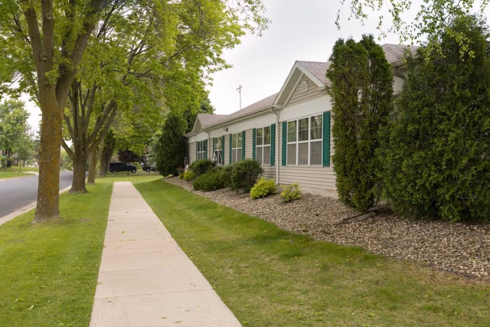 Side view of a single-story building with beige siding and green shutters, surrounded by landscaped bushes and trees, with a sidewalk and grassy area running alongside it.