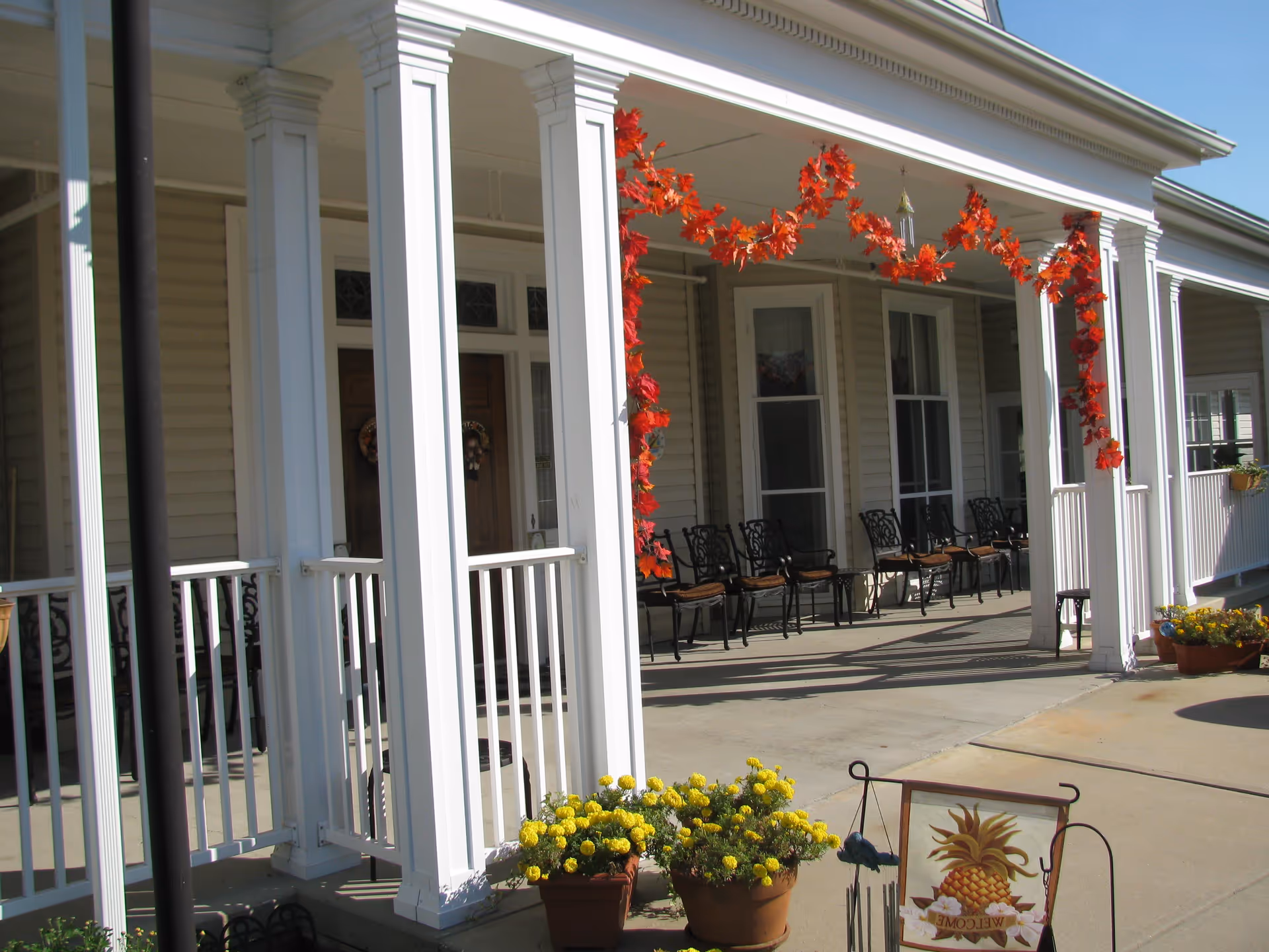 Front porch area of Ridgeway Village Assisted Living with white columns, black metal chairs with cushions, autumn-themed orange leaf garland hanging from the ceiling, and potted yellow flowers near the entrance. A decorative welcome sign with a pineapple is also visible on the ground.