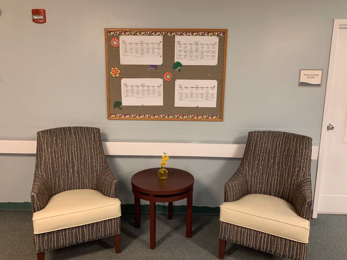 Two patterned armchairs with cream-colored cushions are placed on either side of a small round wooden table with a small yellow flower arrangement in a vase. Behind the chairs is a bulletin board with several papers pinned to it, and to the right is a door labeled 'Medication Room'. The walls are painted light blue and there is a fire alarm on the upper left side.