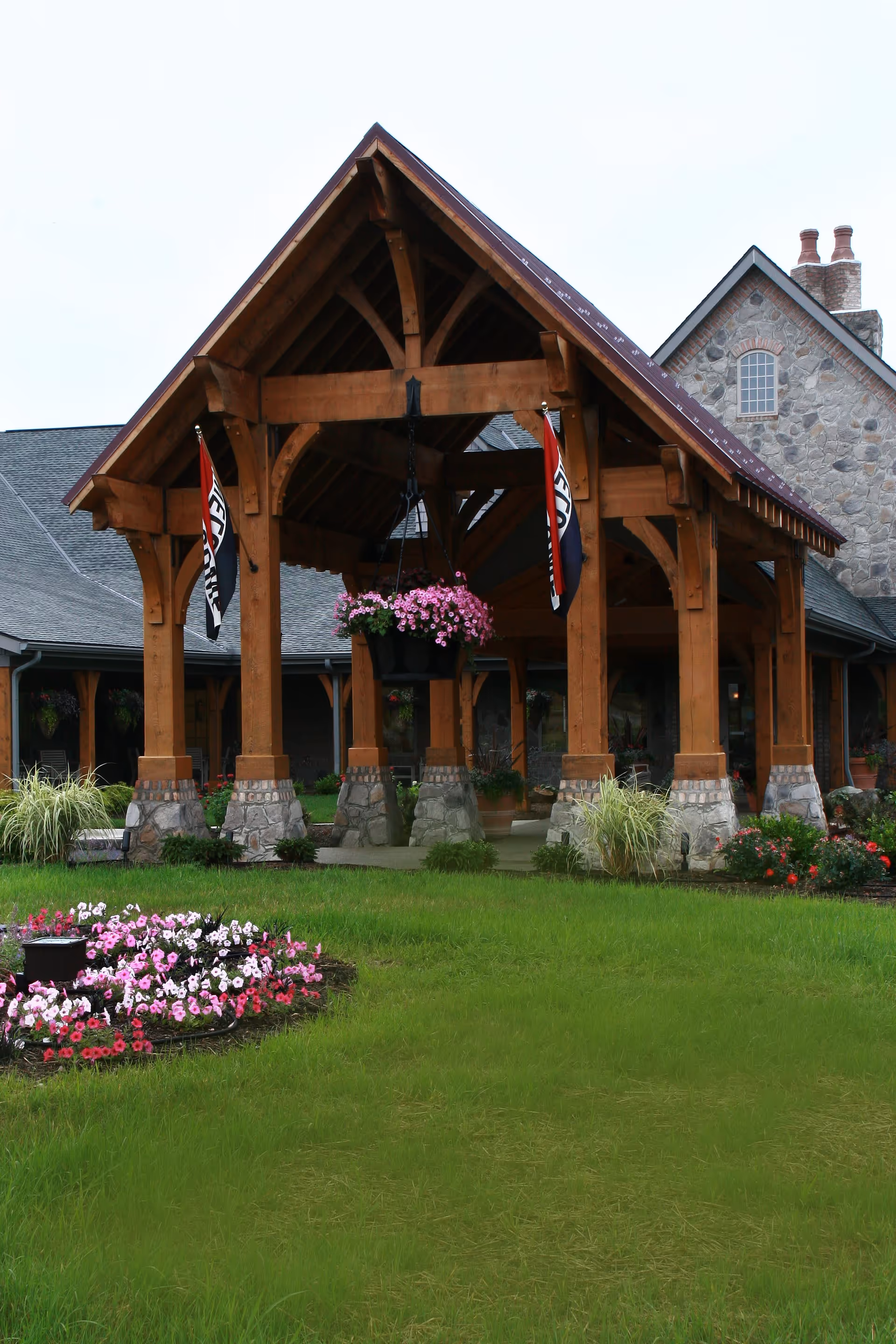 A wooden pavilion structure with stone pillars in front of a stone building with a chimney. The pavilion has hanging flower baskets and two flags. There is a green lawn with flower beds in the foreground.