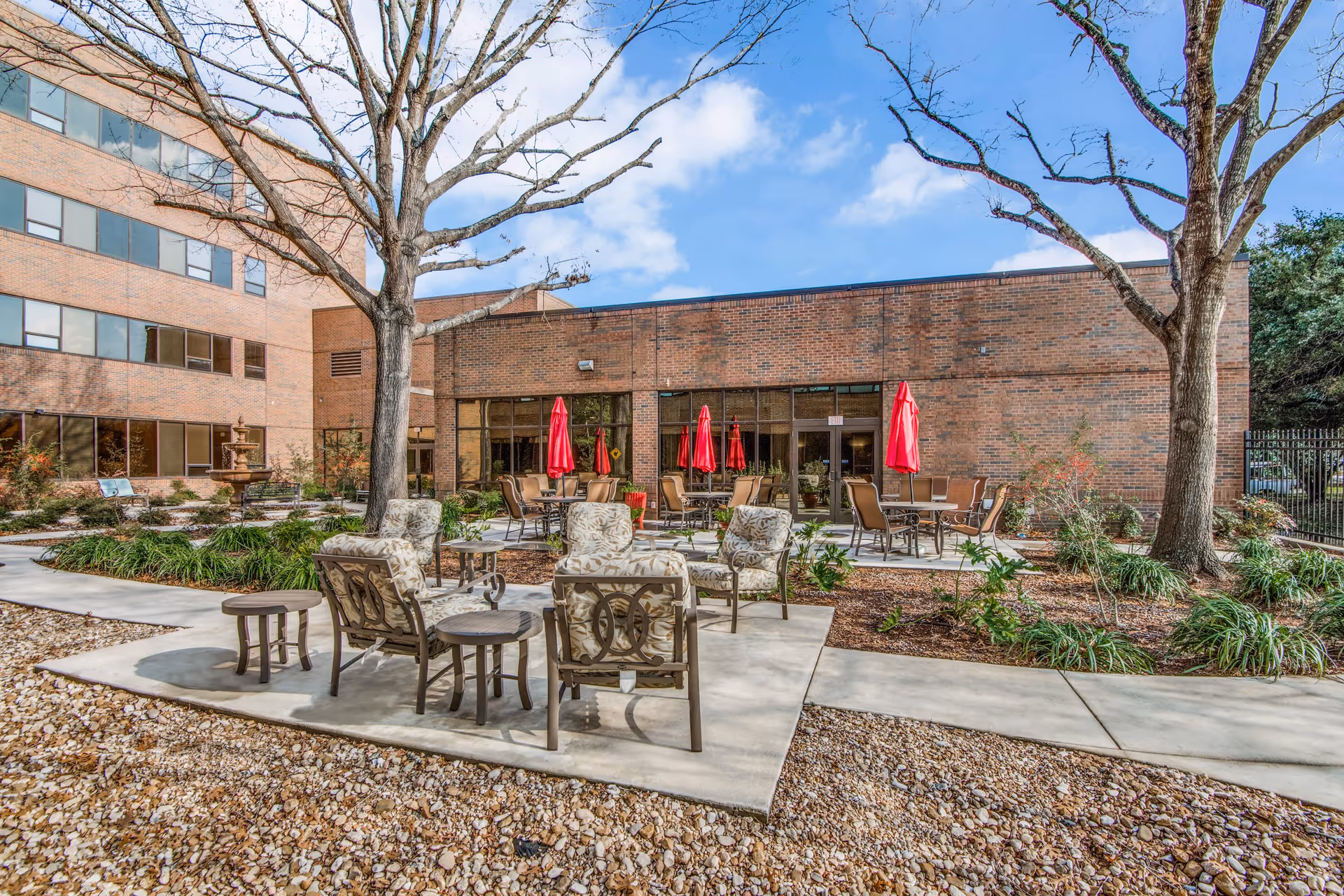 Outdoor patio area at The Heritage at Hunters Chase featuring several cushioned chairs and small tables on a concrete surface surrounded by landscaping with trees and plants. In the background, there are additional tables with red umbrellas and a brick building under a partly cloudy blue sky.