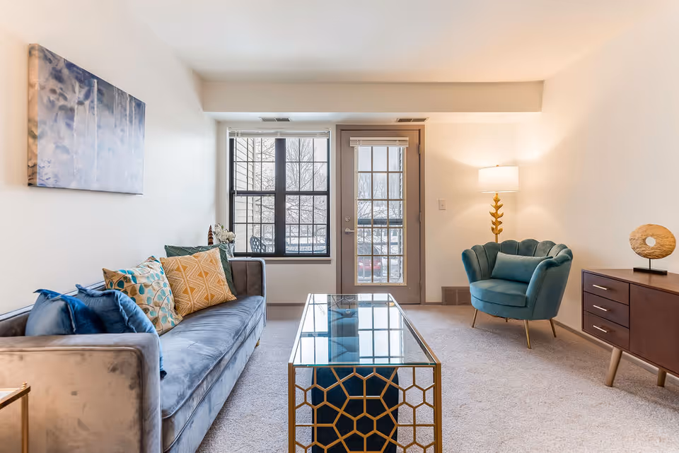 A bright living room with a gray velvet sofa adorned with colorful pillows, a glass coffee table with a gold geometric frame, a teal armchair, a wooden sideboard with decorative items, a floor lamp with a gold base, and a window and glass door letting in natural light.