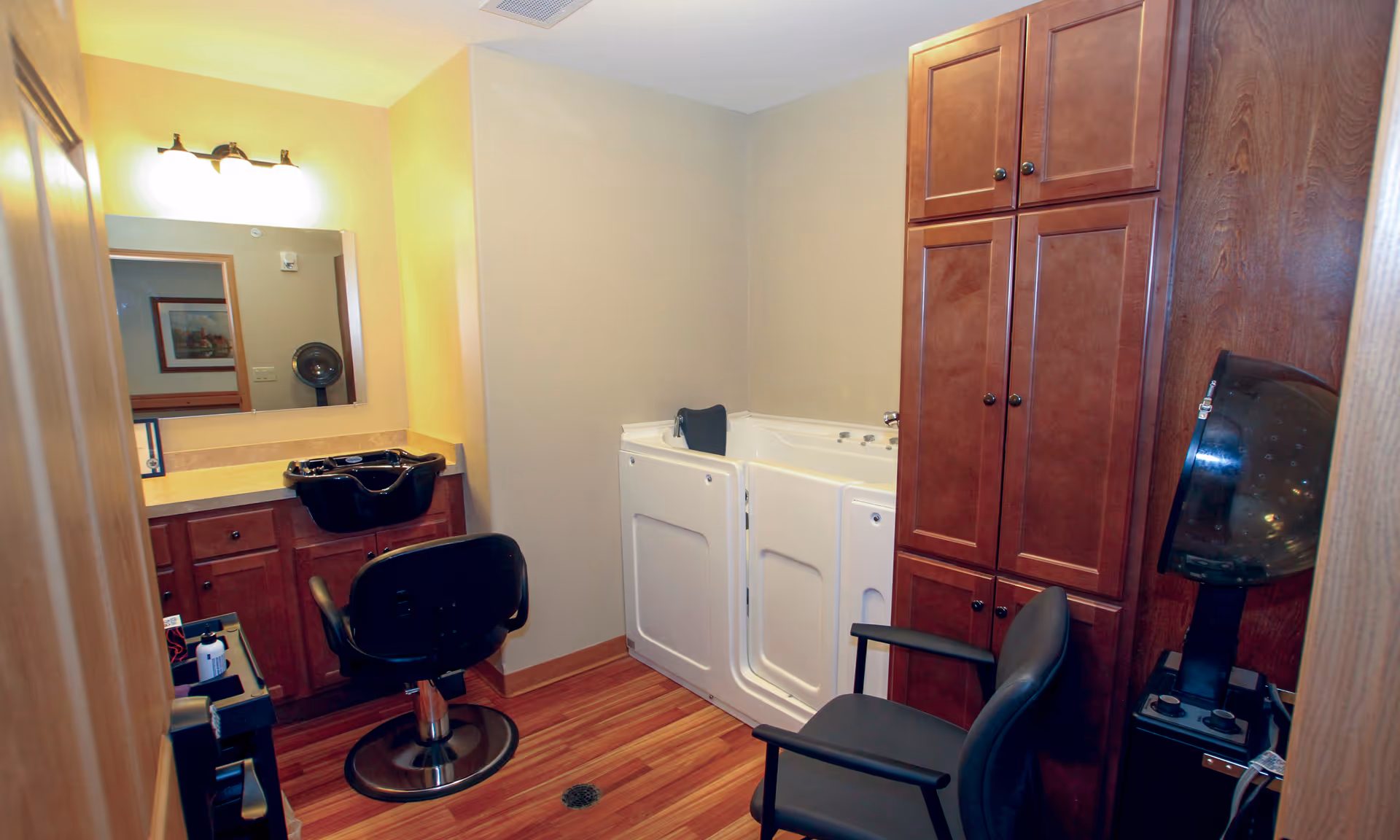 Interior view of a salon room in a senior living facility featuring a black salon chair in front of a sink with a mirror above it, a walk-in bathtub with a seat, wooden cabinets, and a hair dryer hood on the right side.