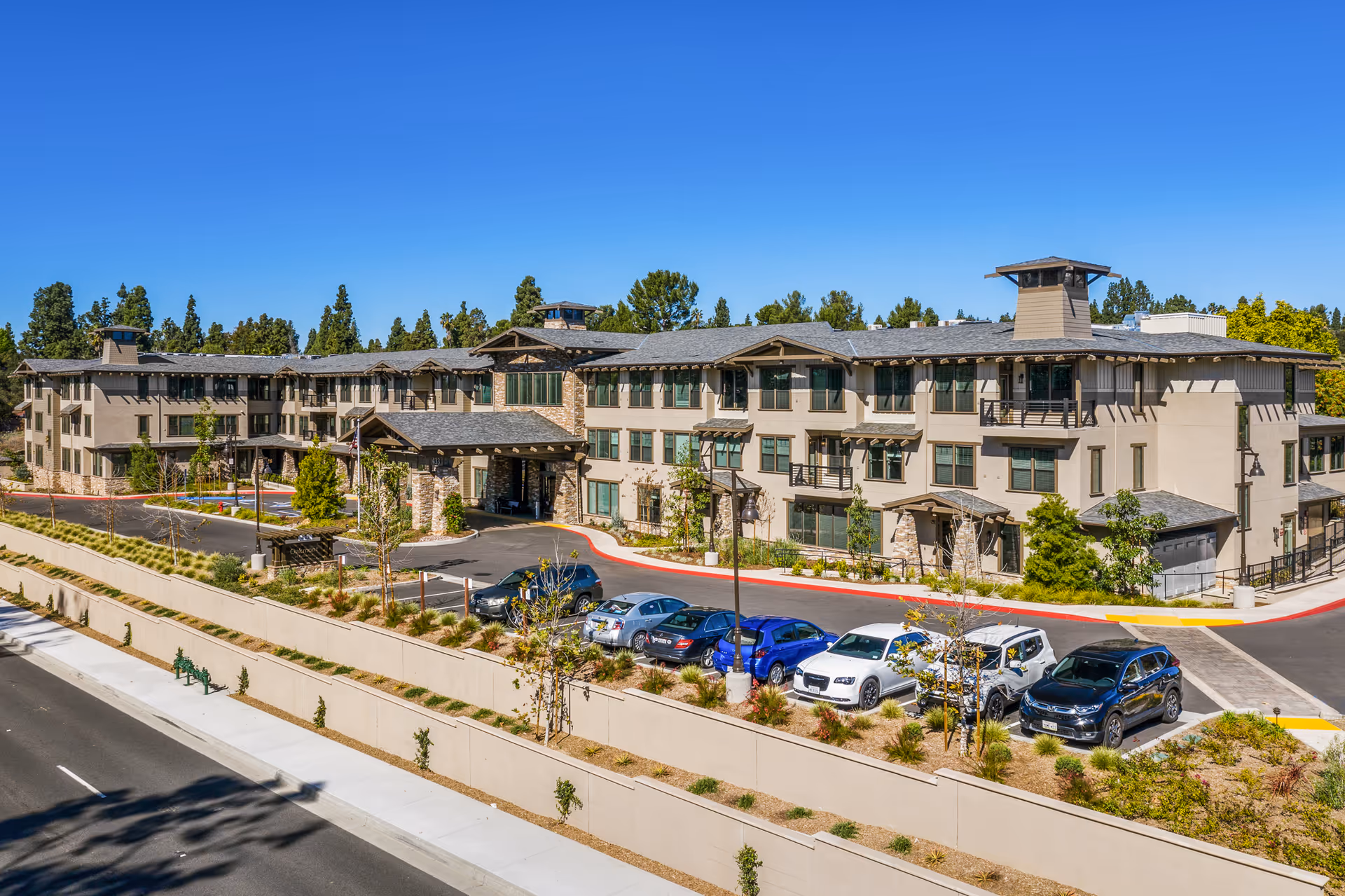 Exterior view of a large, modern three-story senior living facility building with a stone and beige facade, multiple windows, and a covered entrance. Several cars are parked in front of the building, and there are trees and landscaping around the property under a clear blue sky.