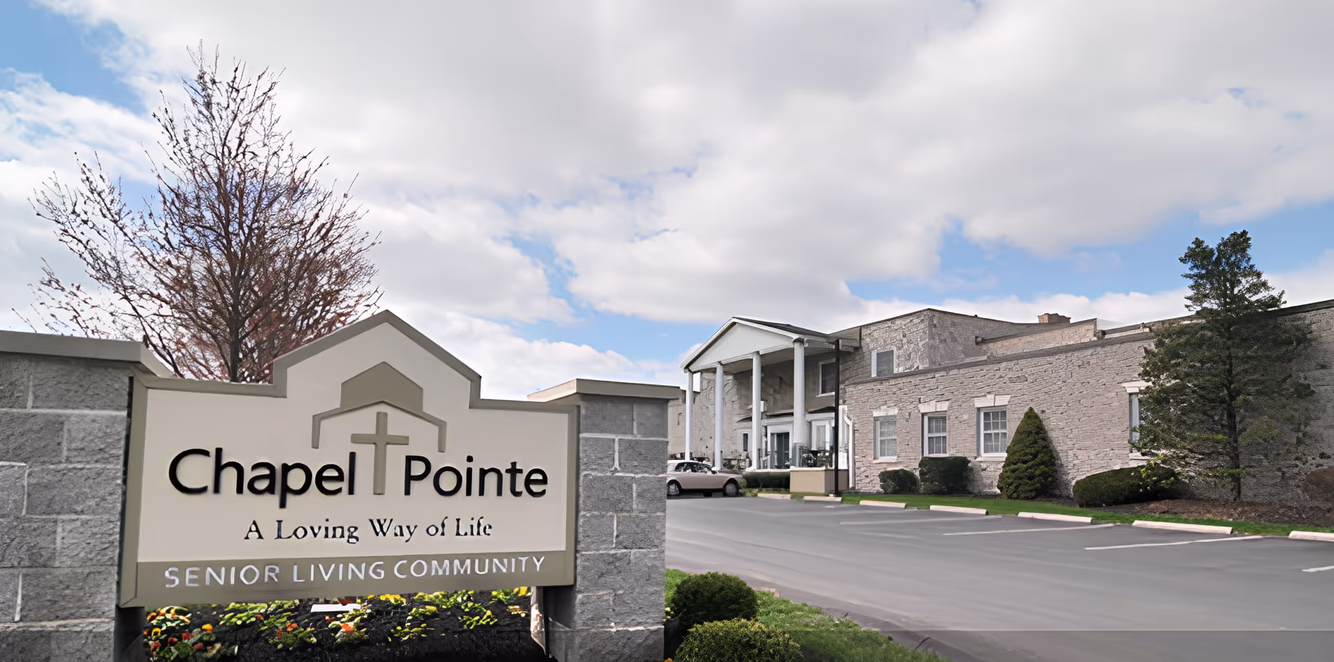 Exterior view of Chapel Pointe At Carlisle senior living community showing a large stone sign with the facility name and slogan, a parking lot, and a building with columns and windows under a partly cloudy sky.