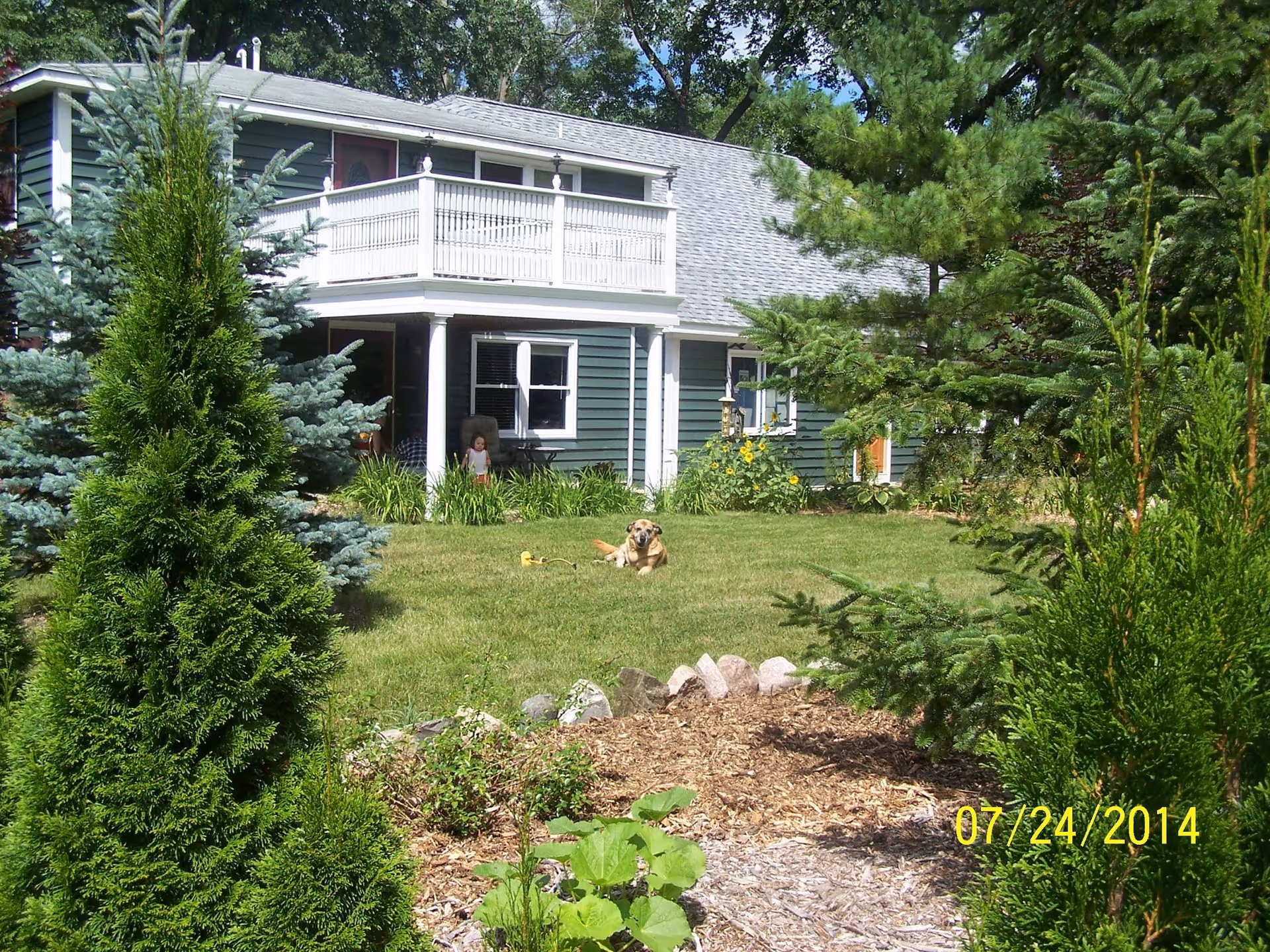 A green two-story house with a balcony and white pillars surrounded by trees and shrubs. A dog is lying on the grass in the front yard. The date 07/24/2014 is visible in the bottom right corner.