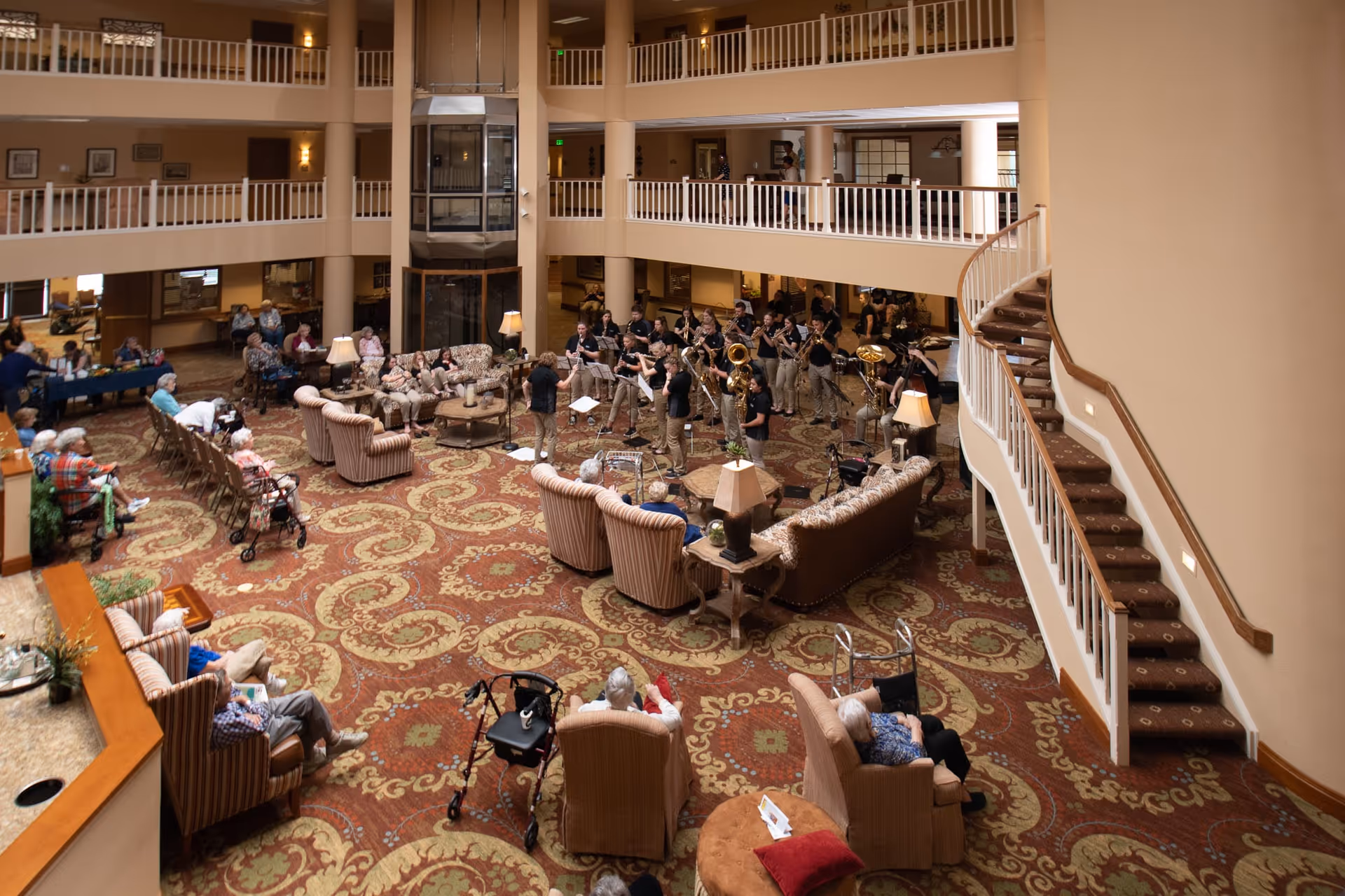 A spacious, multi-level common area in a retirement village with patterned carpet, several armchairs and sofas arranged around coffee tables, and a group of musicians performing in the center. Elderly residents are seated around the room watching the performance. A staircase with a carpeted runner leads to the upper floors with railings overlooking the area.