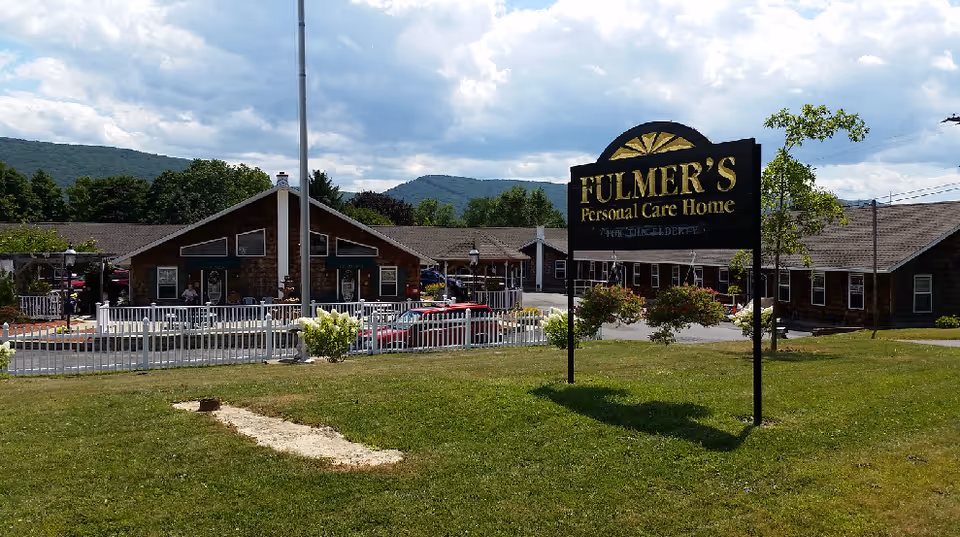 Exterior view of Fulmer's Personal Care Home showing a large sign with the facility's name on a grassy lawn. The building is a single-story structure with a brown roof and multiple windows. There is a white picket fence and some outdoor seating visible near the entrance. Trees and hills are in the background under a partly cloudy sky.