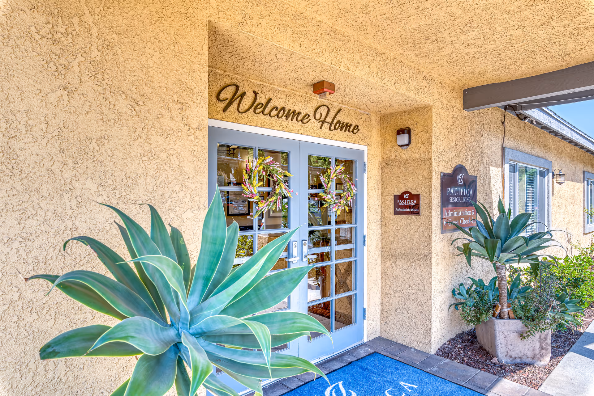 Entrance to a senior living facility with double glass doors decorated with floral wreaths. Above the doors is a sign that reads 'Welcome Home'. There are large potted plants on either side of the entrance and signs indicating 'Pacifica Senior Living Administration & Guest Check-In'. The building exterior is textured beige stucco.