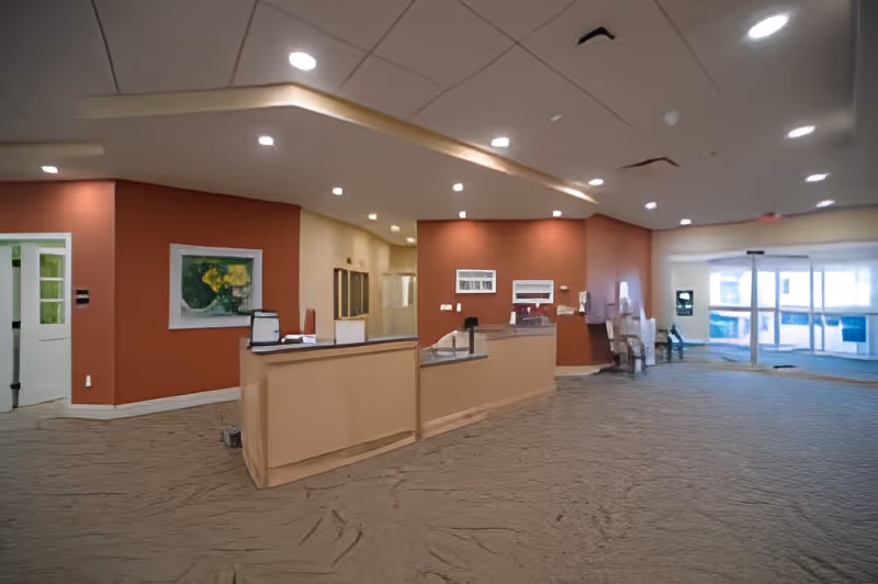 Reception area inside a senior living facility with a beige front desk, red and beige walls, recessed ceiling lights, a painting on the wall, and large glass doors leading outside.