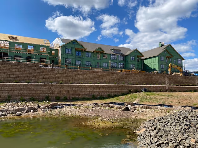 Exterior view of a large green-clad building under construction behind terraced stone retaining walls and a small pond.