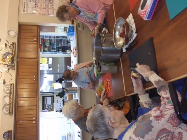 Four elderly women are gathered around a kitchen table preparing food. One woman is cutting vegetables on a red cutting board, while another is peeling or cutting fruit. There are large pots and bowls on the table, and the kitchen cabinets and appliances are visible in the background.