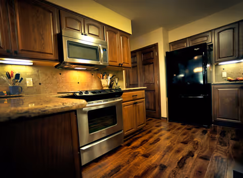A kitchen with wooden cabinets, a stainless steel oven and microwave, a black refrigerator, and wooden flooring. The countertops are granite, and there are under-cabinet lights illuminating the backsplash and workspace.