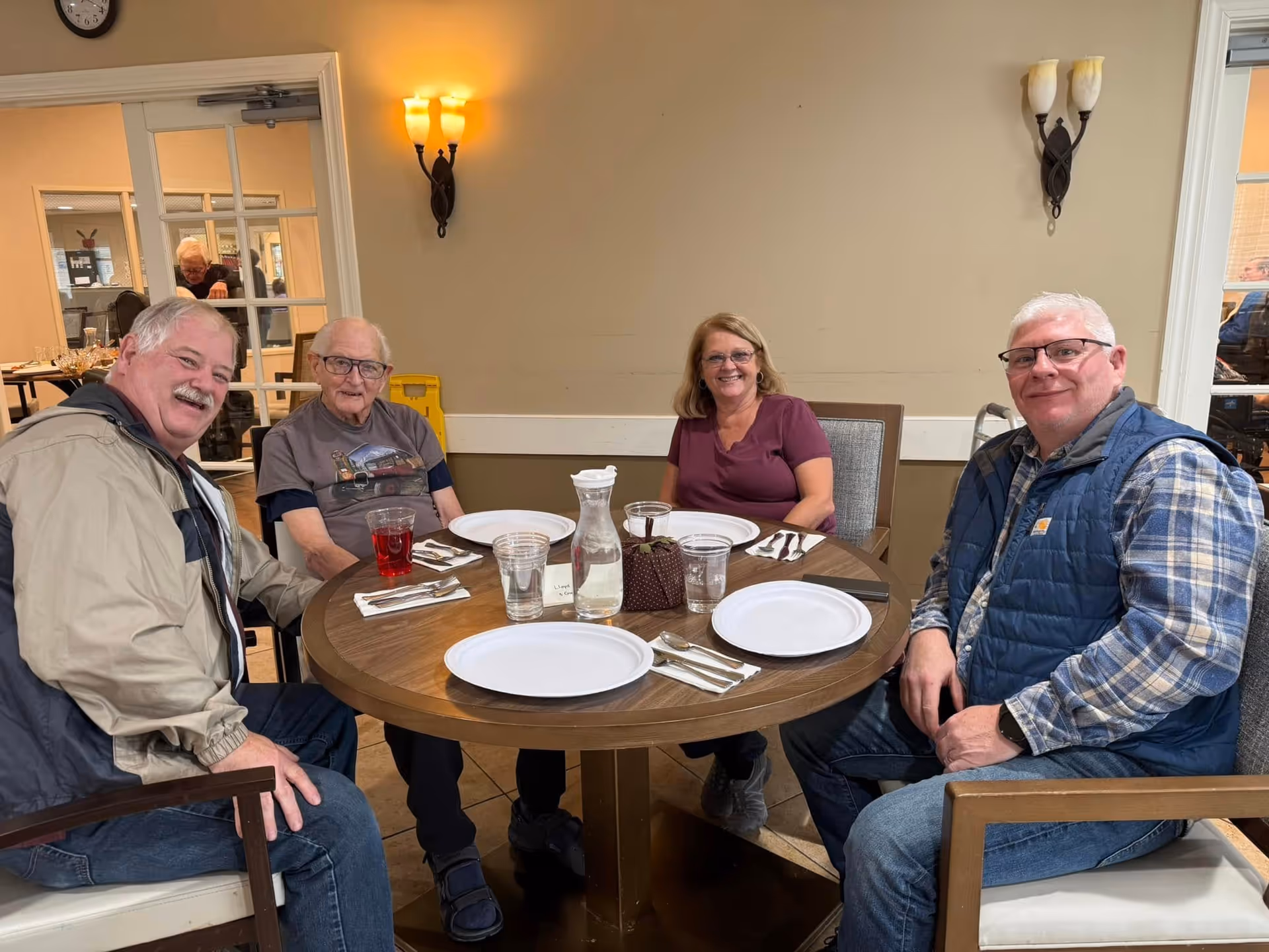 Four adults sitting around a round wooden dining table set with empty white plates, utensils, glasses of water, and a pitcher of water in a well-lit room with beige walls and wall-mounted light fixtures.