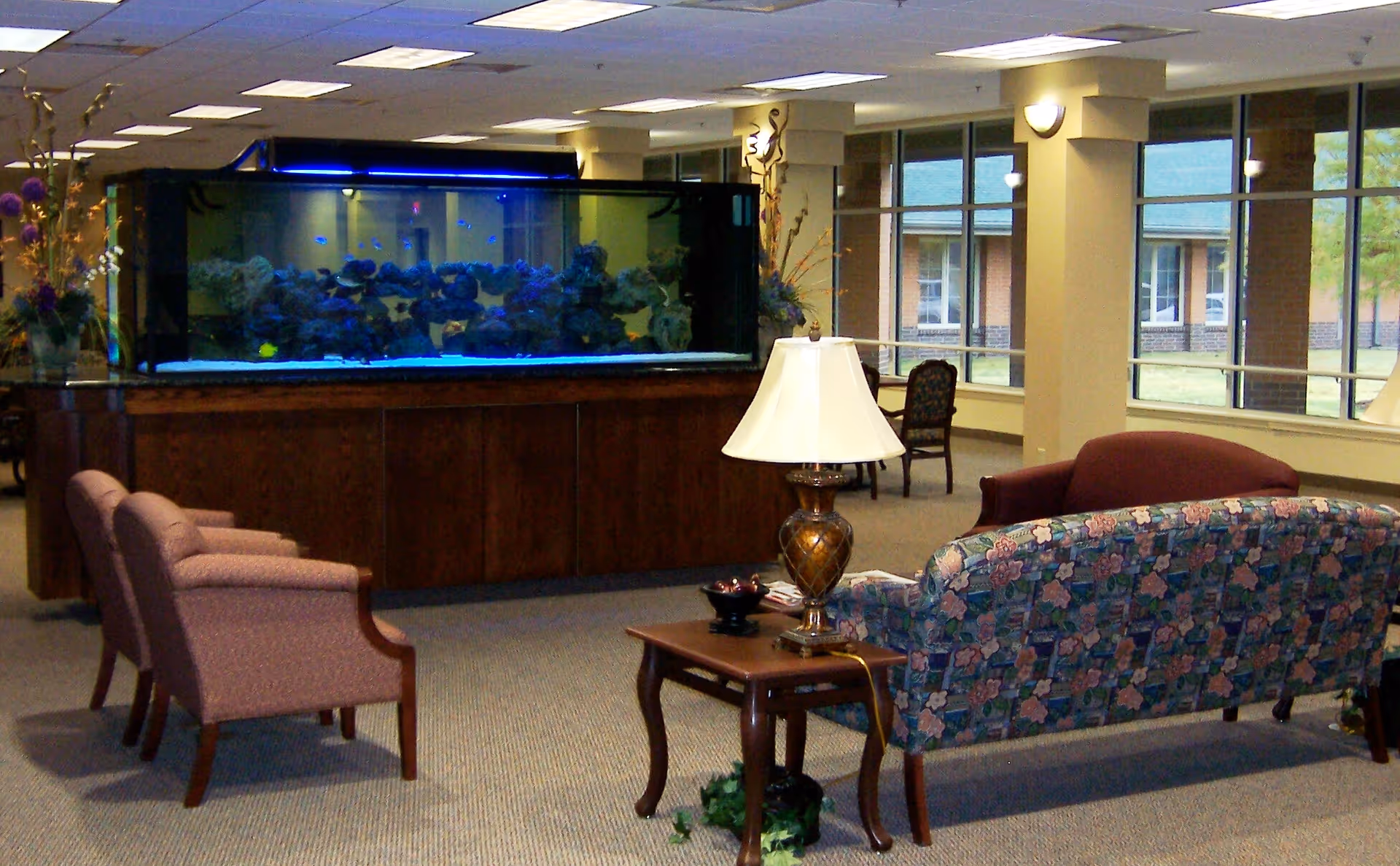 A spacious common area with a large aquarium filled with fish on a wooden stand. The room has several upholstered chairs and sofas arranged around a wooden side table with a decorative lamp. Large windows allow natural light to enter, and there are decorative plants near the aquarium.