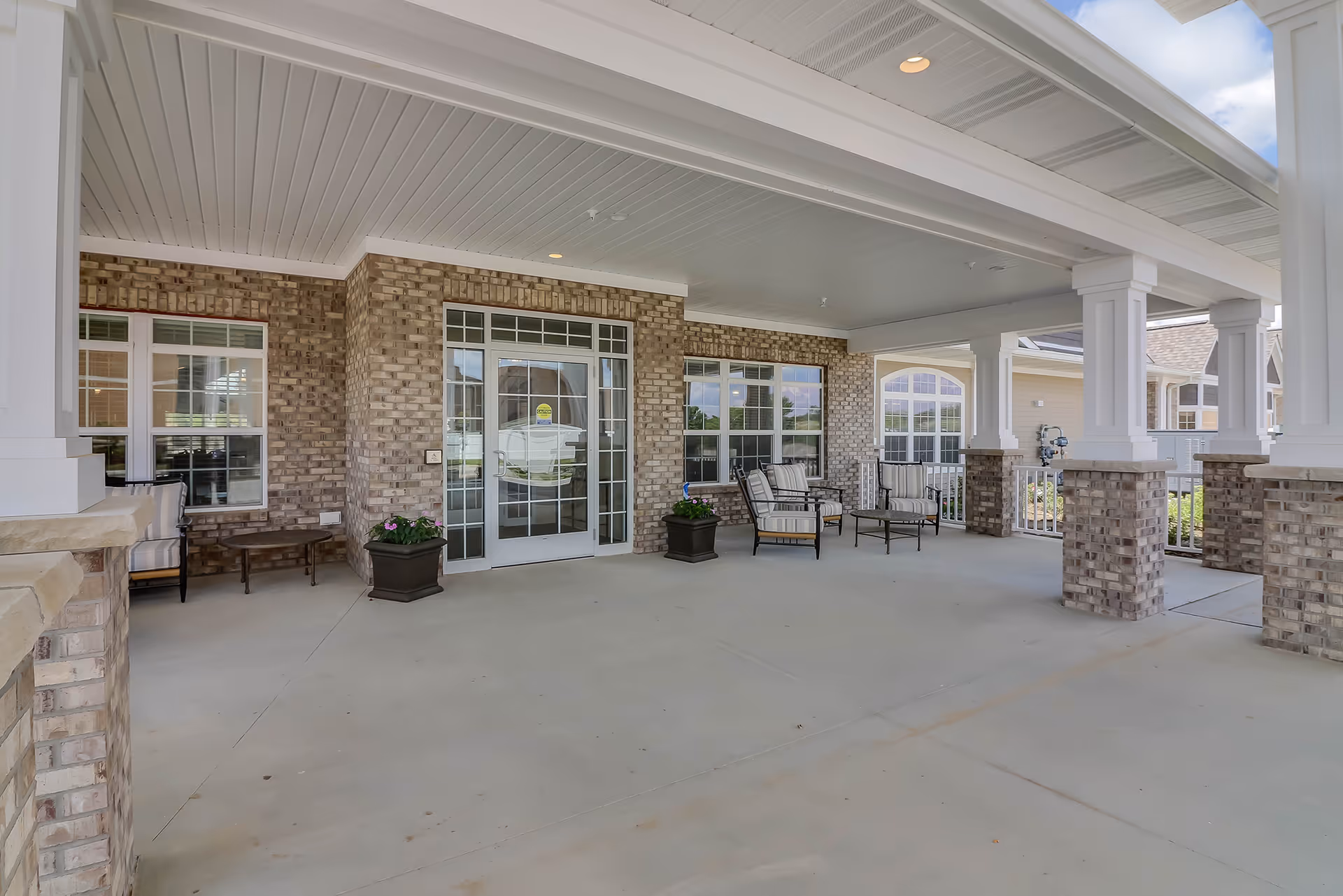 Covered brick front entrance with white columns, outdoor seating, planters, and glass double doors.